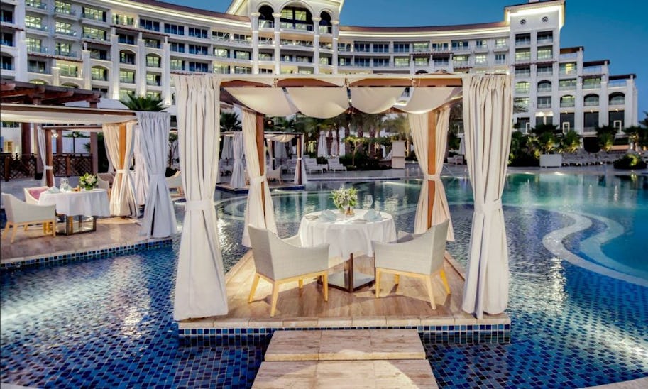 A table for two set up in a pergola that's sitting out on a pontoon the middle of a swimming pool